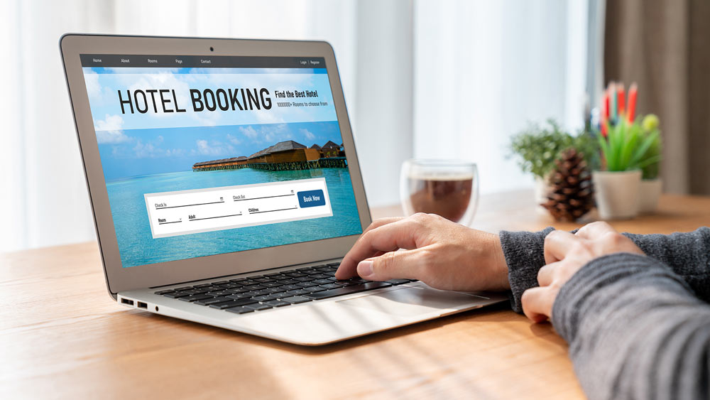 Auf dem Bild ist ein Laptop zu sehen, auf dessen Bildschirm eine Webseite für Hotelbuchungen geöffnet ist. Die Seite zeigt das Wort "HOTEL BOOKING" in großen Buchstaben und eine idyllische Szenerie eines Resorts am Meer, mit Bungalows, die auf Stelzen über das Wasser gebaut sind. Darunter befindet sich ein Formular zur Eingabe von Daten für Check-in und Check-out, Zimmeranzahl sowie Angaben zu Erwachsenen und Kindern.